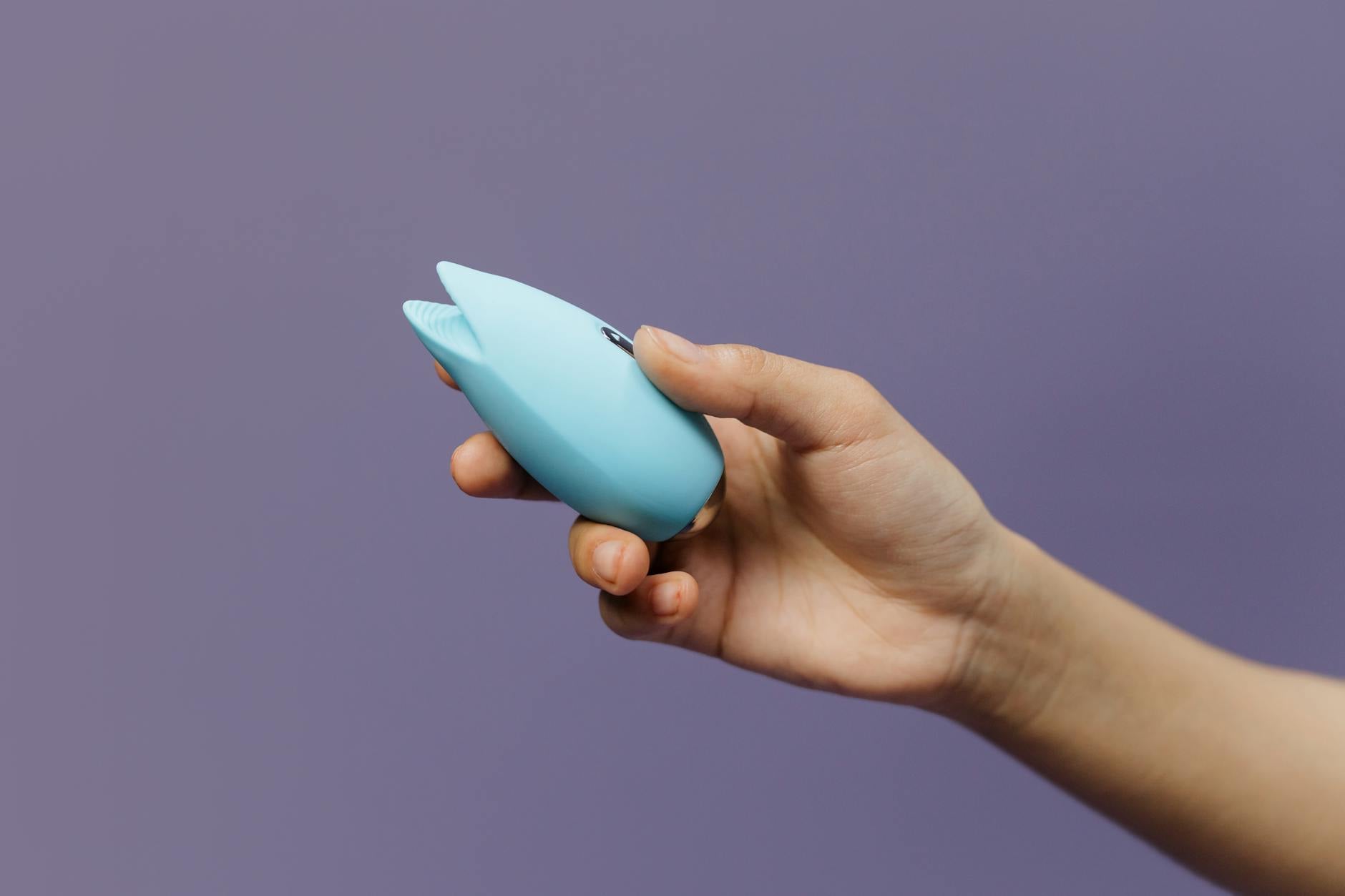 A hand holding a blue silicone vibrator against a purple background, showing texture detail