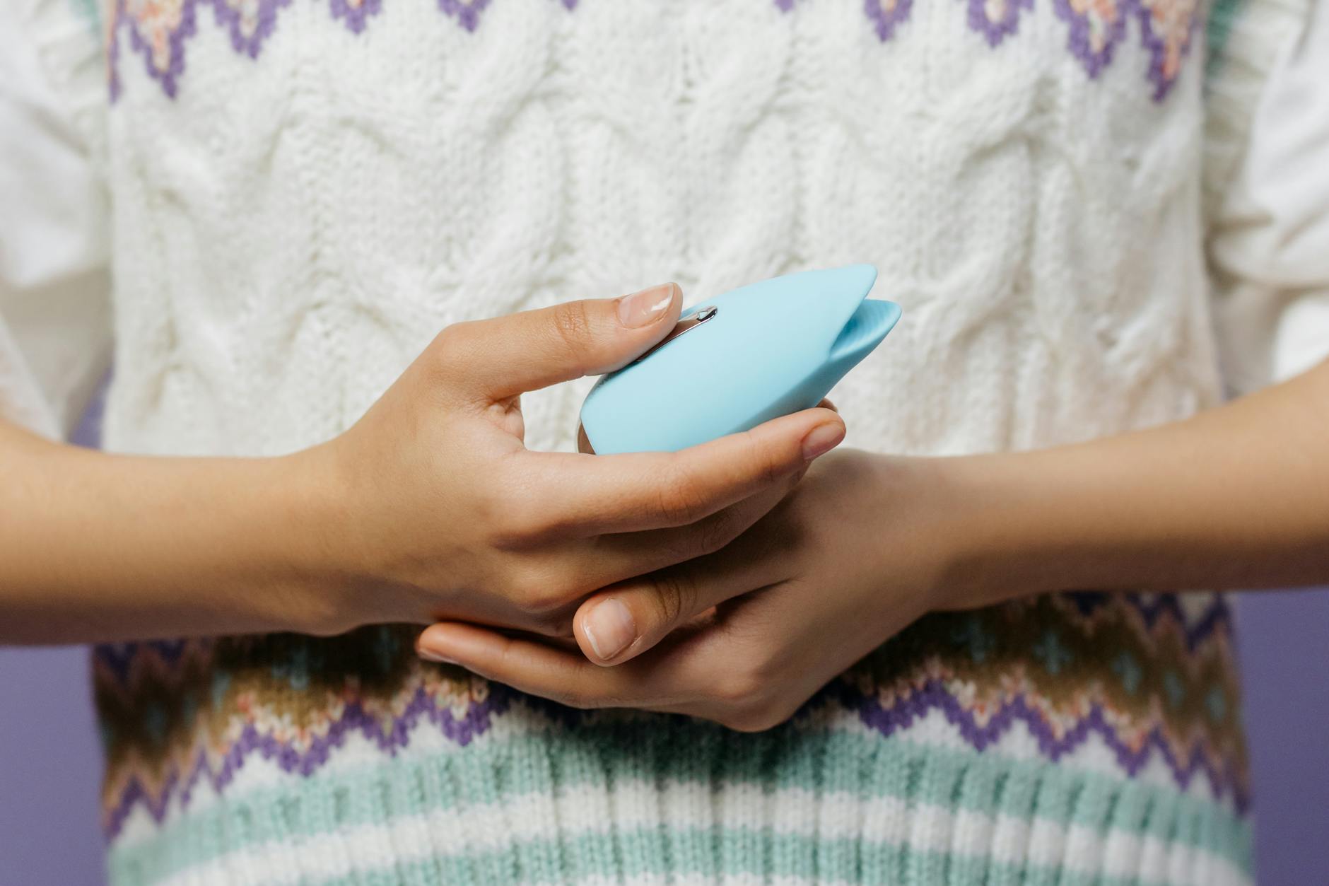 Hands holding a blue vibrator against a cozy knitted sweater, representing comfort and intimate self-care.